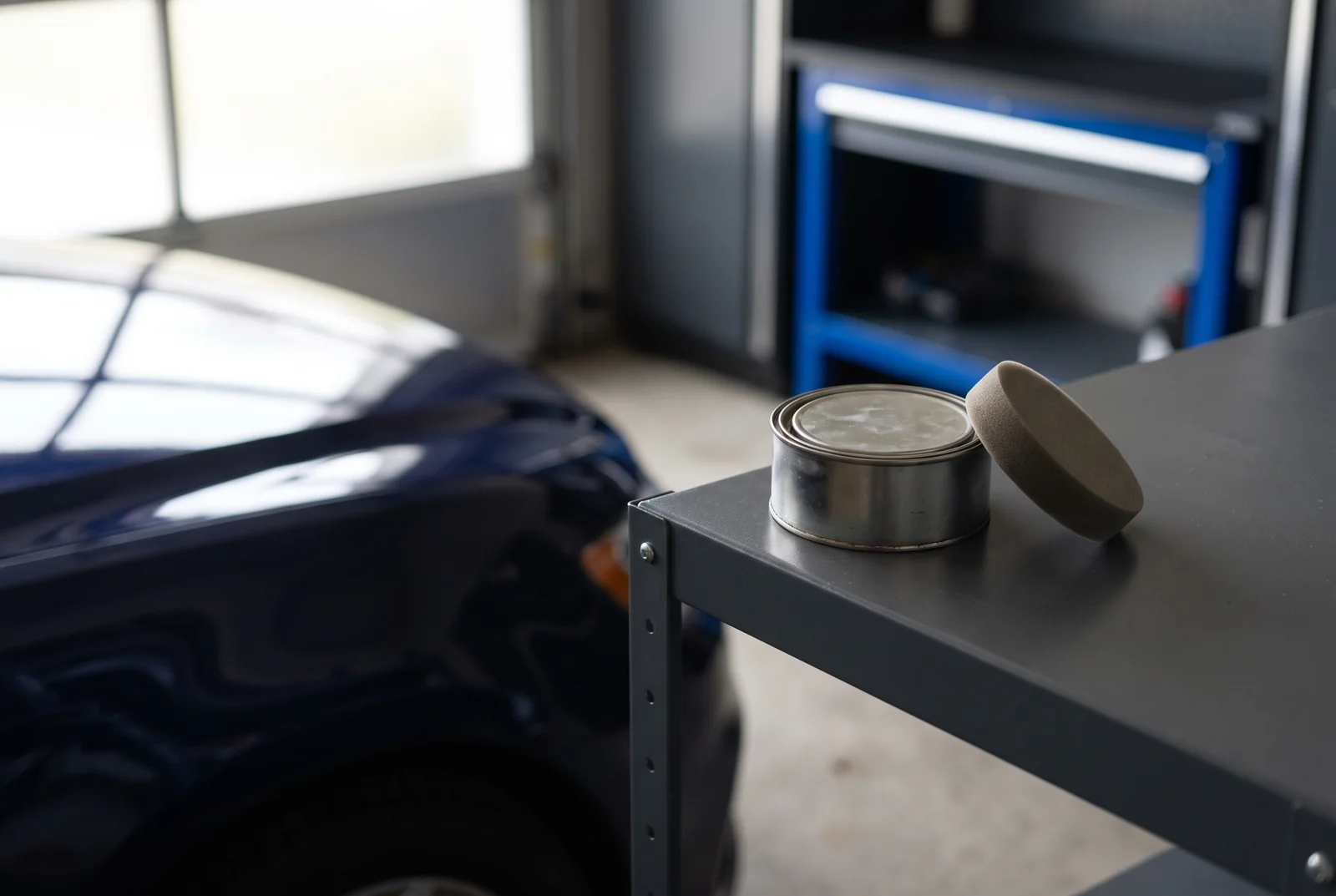 A wax tin and foam applicator on a shelf beside a navy car.
